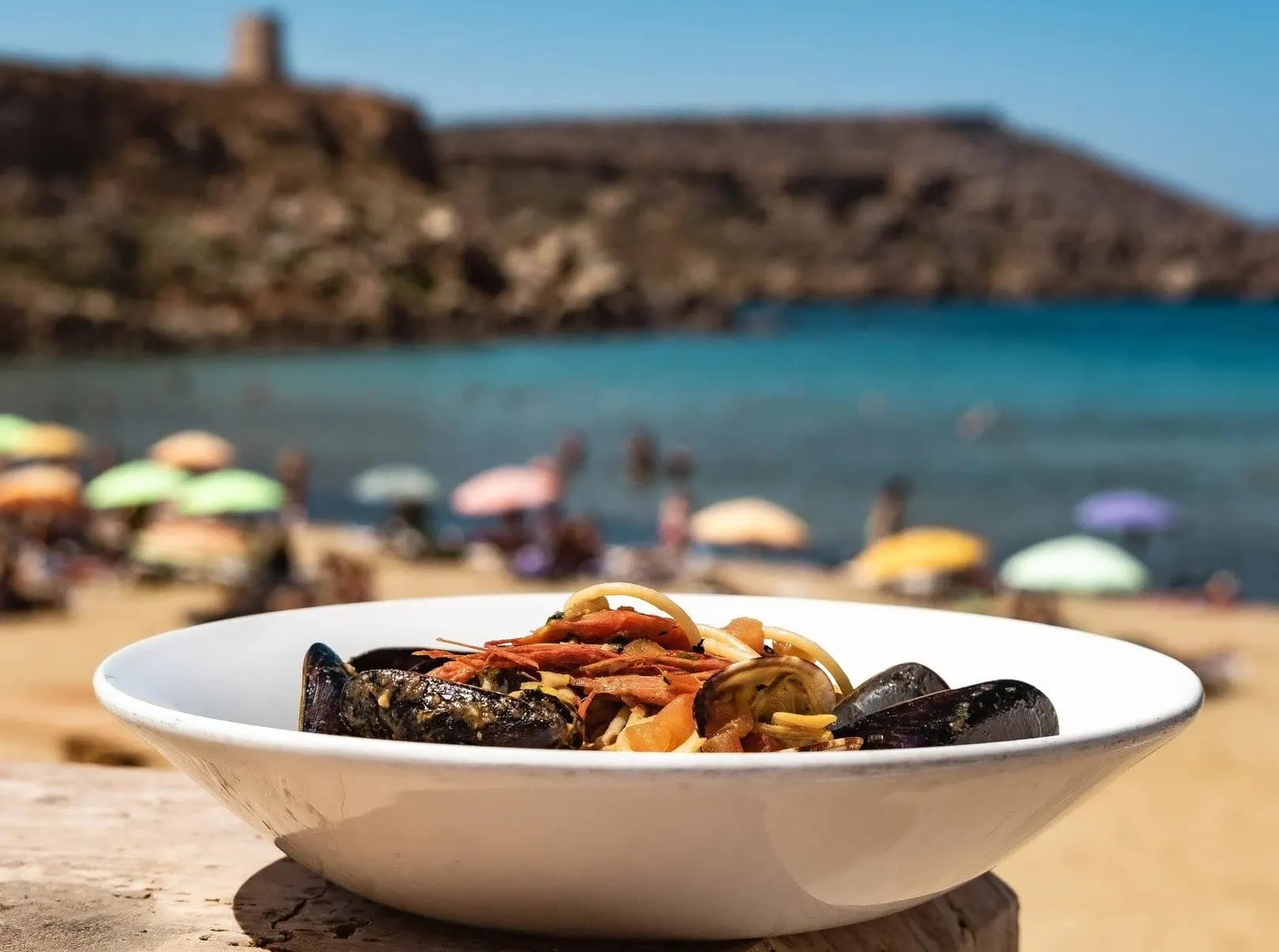 A shot of a plate of seafood pasta, with sand, sea and architecture in the background. A typical site on a Malta holiday.